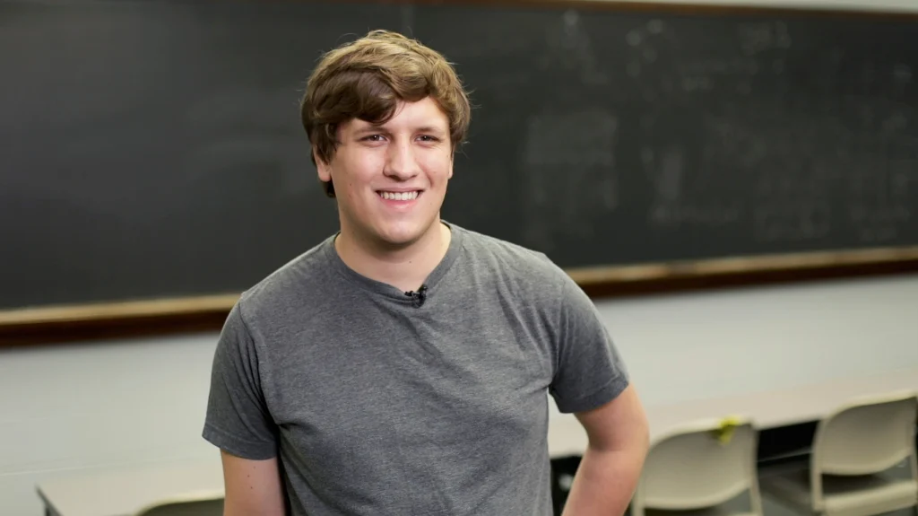A smiling young man in a gray t-shirt stands in a classroom with a chalkboard and empty chairs, ready to inspire others to take research beyond the lab.