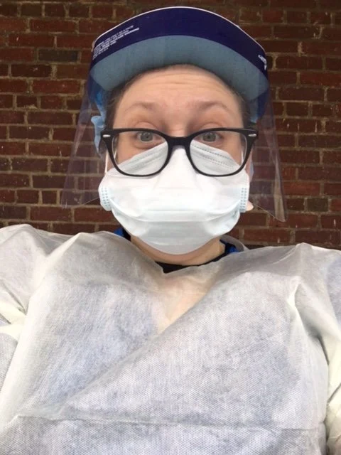 Person wearing a face shield, glasses, mask, and protective gown stands on the front lines in front of a brick wall.