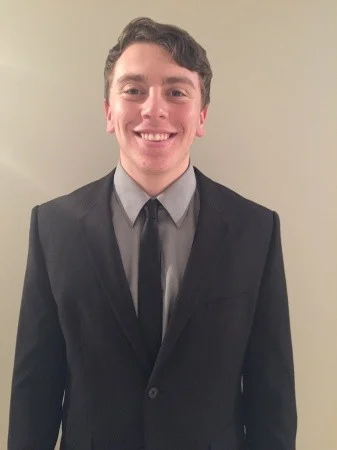 A young man in a black suit, gray shirt, and black tie—one of many proud public health graduates—smiles in front of a plain light-colored wall.