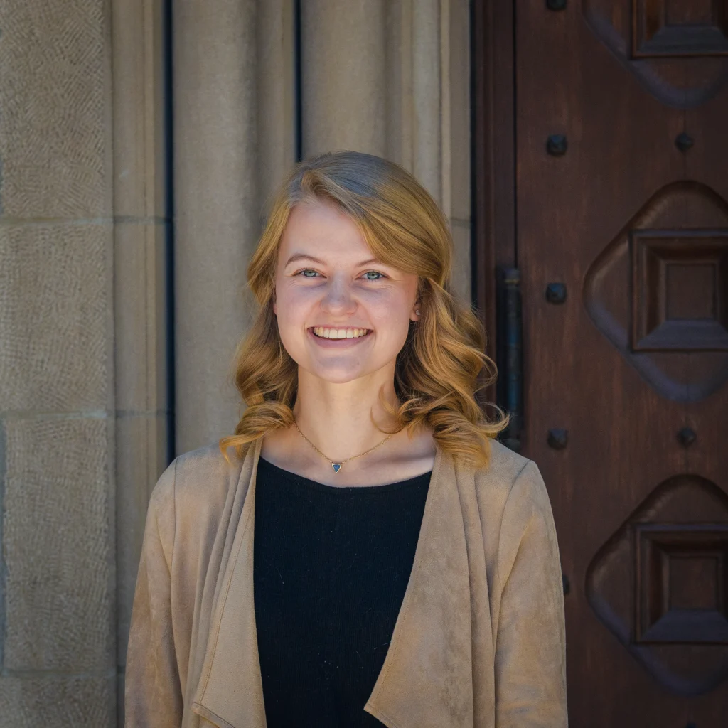 A young woman with blonde hair smiles, standing in front of a stone and wooden doorway, her expression radiating quiet faith.