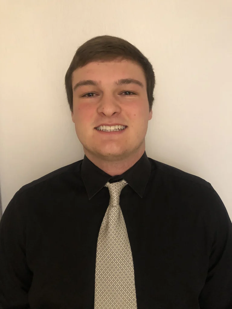 Young man in a black shirt and light-colored tie smiling in front of a plain light background, reflecting his pursuit of quality in both style and presentation.