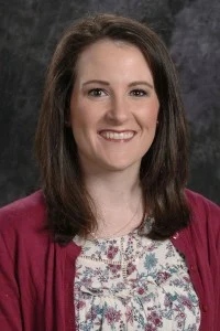 Woman with shoulder-length brown hair wearing a floral blouse and maroon cardigan, smiling at the camera, radiating warmth and occupational ambition.