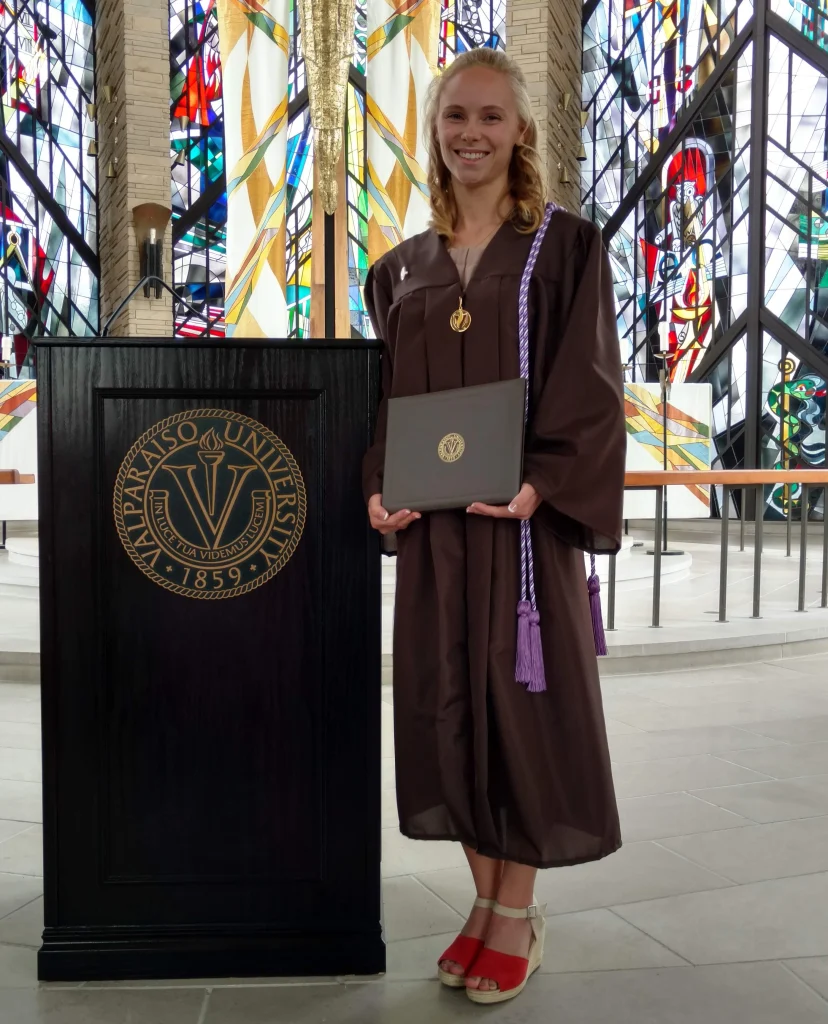 Young woman in a brown graduation gown holding a diploma, standing by a podium at Valparaiso University, celebrates her Nursing degree and the powerful connections she’s made along the way.