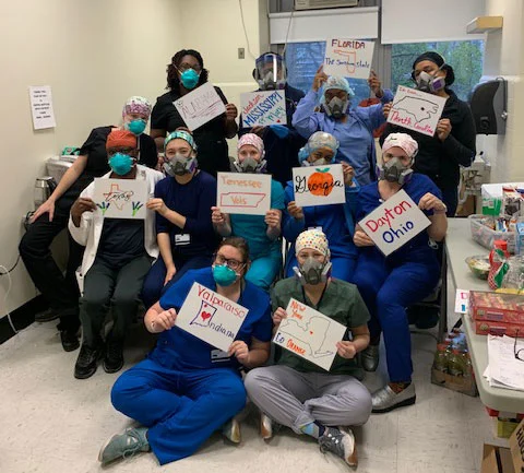 A group of healthcare workers on the front lines in masks hold up signs showing names of U.S. states and cities.