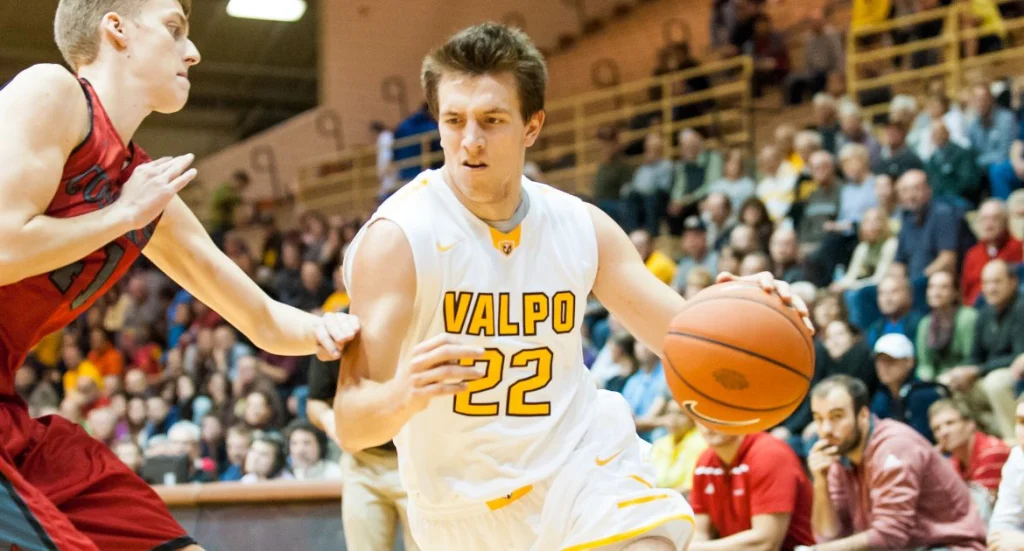 Valpo student-athlete in a white jersey dribbles past a defender mid-game, with a lively crowd in the background, honoring frontline workers and their efforts during COVID-19.