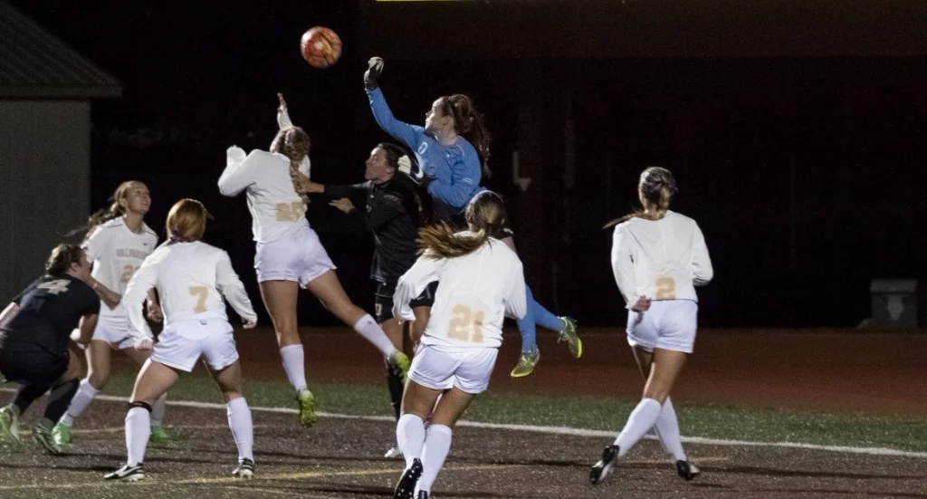 A soccer goalie jumps to punch the ball away as Valpo Student-Athletes in white and black uniforms compete in the box at night.