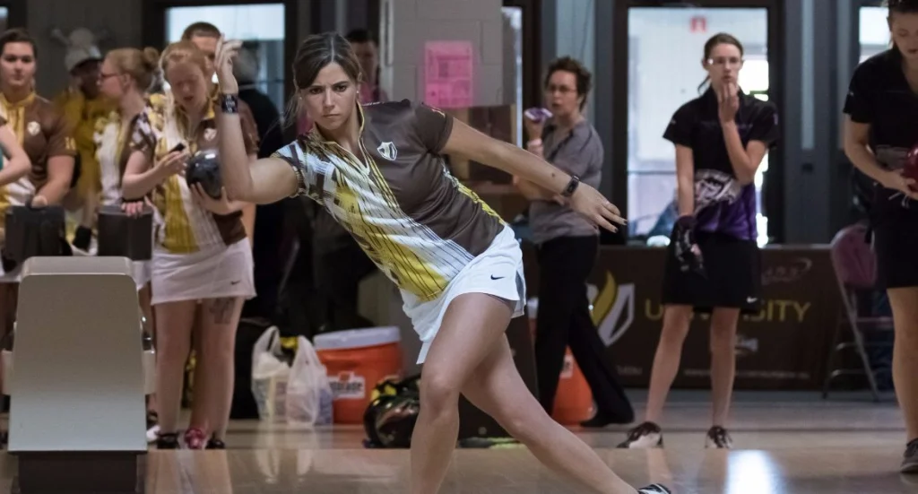 A woman in sports attire, likely one of the Valpo Student-Athletes, bowls at an indoor alley, with other players and spectators social distancing in the background due to COVID-19 protocols.