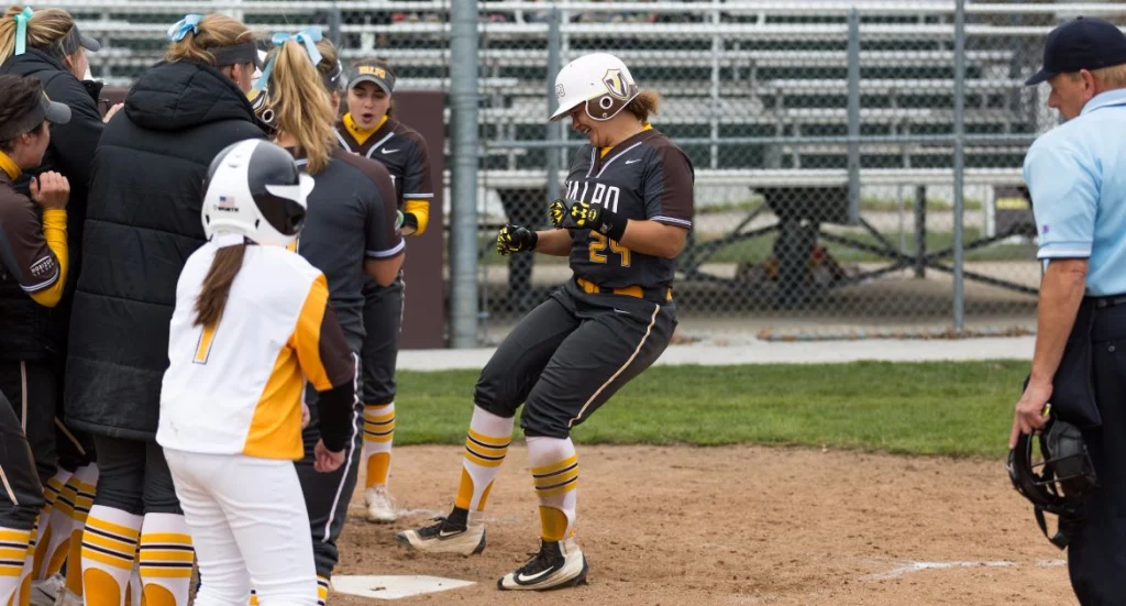 Valpo Student-Athletes celebrate at home plate as teammates cheer near a fence; umpire stands to the right.