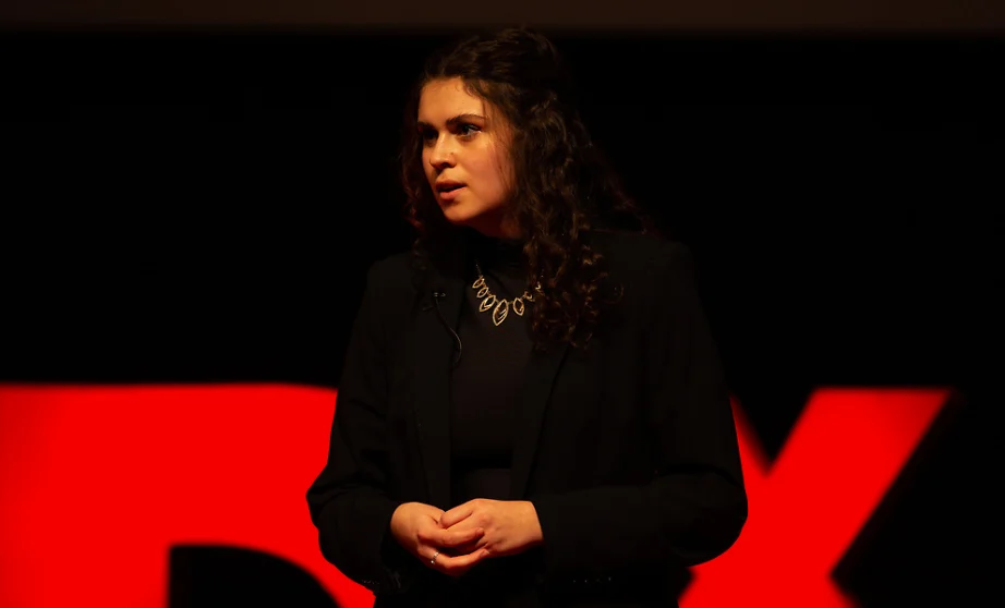 A Creative Writing Major in dark clothing speaks on stage in front of a large red TEDx Valpo sign.