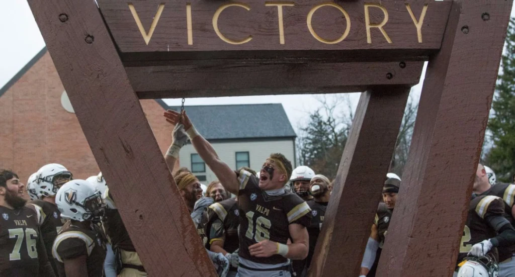 Football player in uniform rings victory bell, surrounded by celebrating Valpo Student-Athletes under a large "VICTORY" sign.