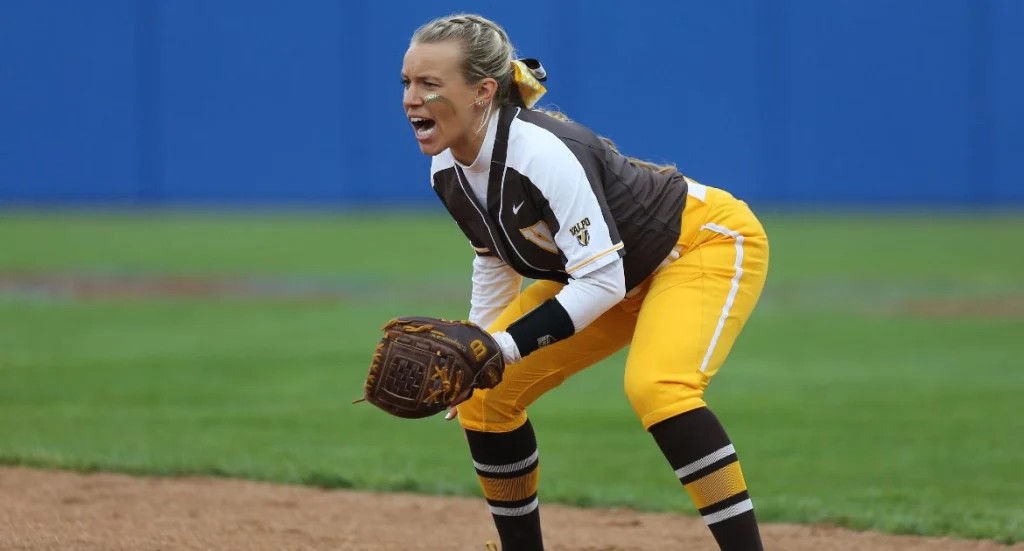 Valpo Student-Athletes: Softball player in yellow and brown uniform shouting while fielding on a dirt infield, glove ready.