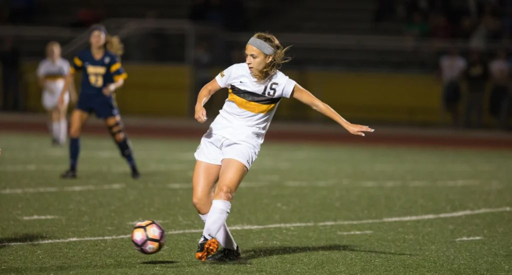 A Valpo student-athlete in a white uniform kicks the ball during a game on a grassy field, showcasing determination reminiscent of frontline workers during COVID-19.