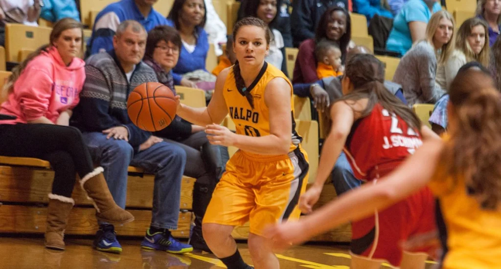 A female Valpo student-athlete in yellow dribbles while looking to pass, with spectators watching in the background, honoring the resilience shown by frontline workers during COVID-19.