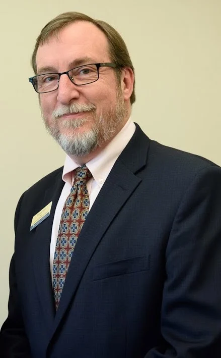 A man in a suit and patterned tie, wearing glasses, stands against a plain light background, smiling slightly—reflecting the professionalism of an Associate Provost for Graduate and Online Education at Valparaiso University.