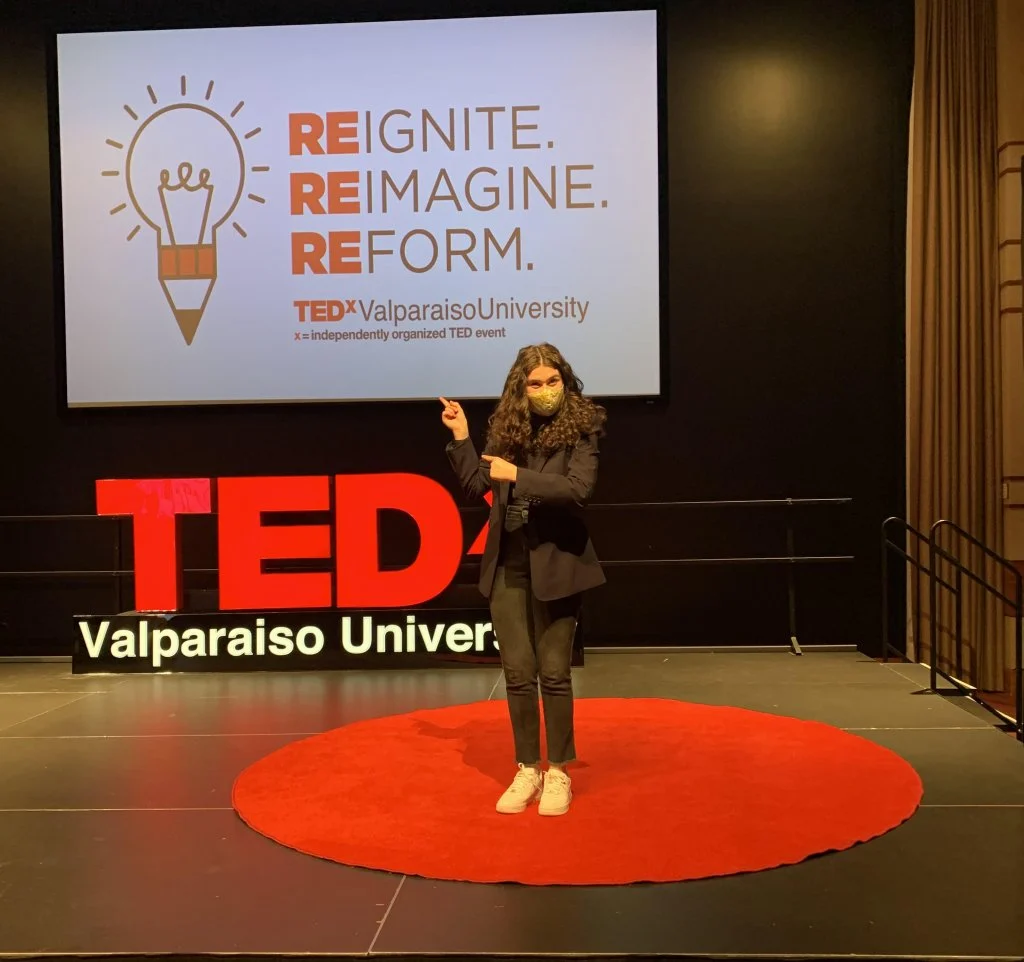 A speaker stands on a red circle stage at Valpo TEDx, pointing at a screen behind them, as Creative Writing Major Abby Seeber shares her insights.