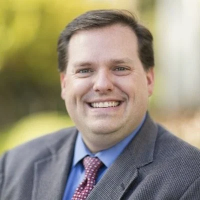 Smiling man in a suit and tie outdoors with a blurred green background, representing Valparaiso University’s Vice President of Enrollment.