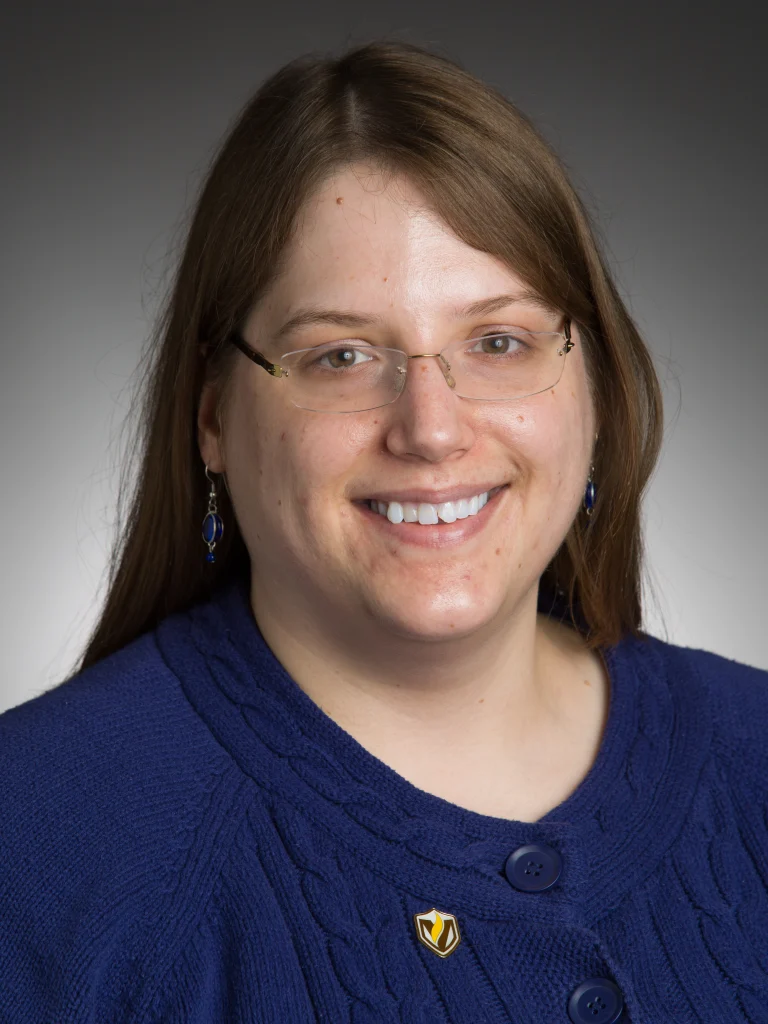 Woman with long brown hair, glasses, and blue sweater smiling at the camera against a gray background, reflecting her role as a Valparaiso University professor known for excellence in teaching.