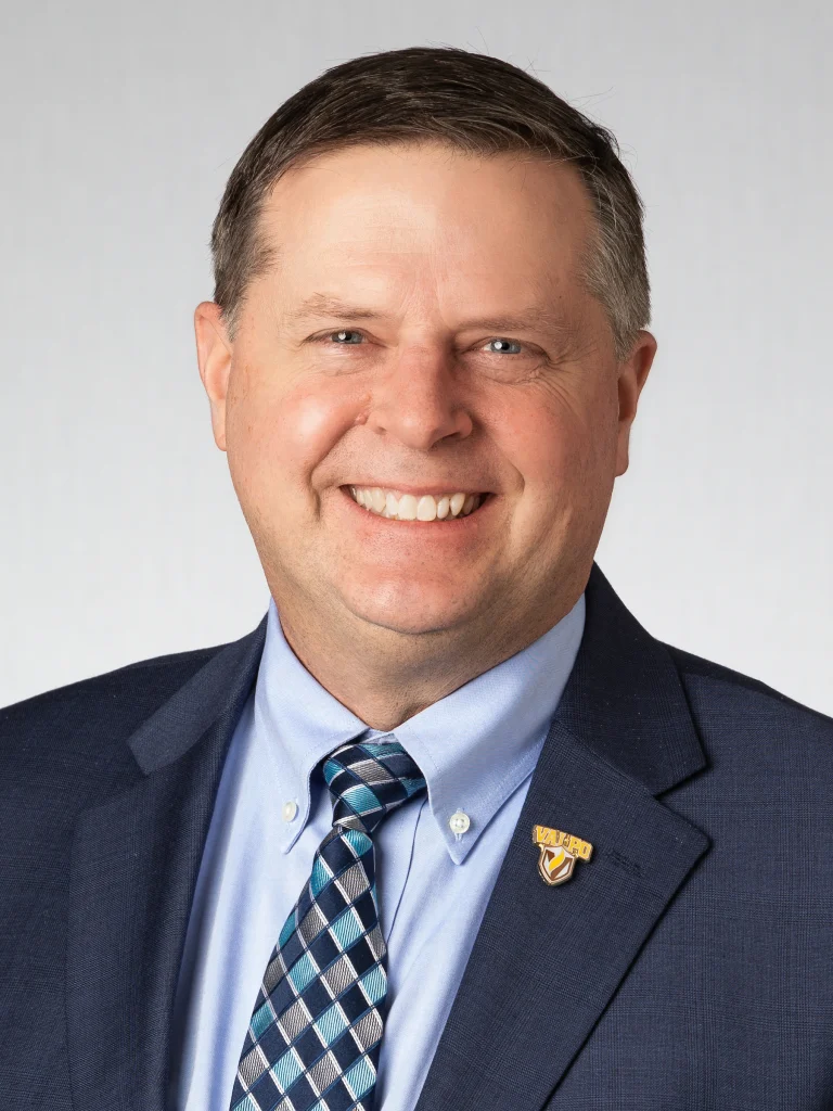 Smiling man in a suit and tie with short brown hair, serving as Provost and Executive Vice President for Academic Affairs at Valparaiso University, poses against a light gray background.
