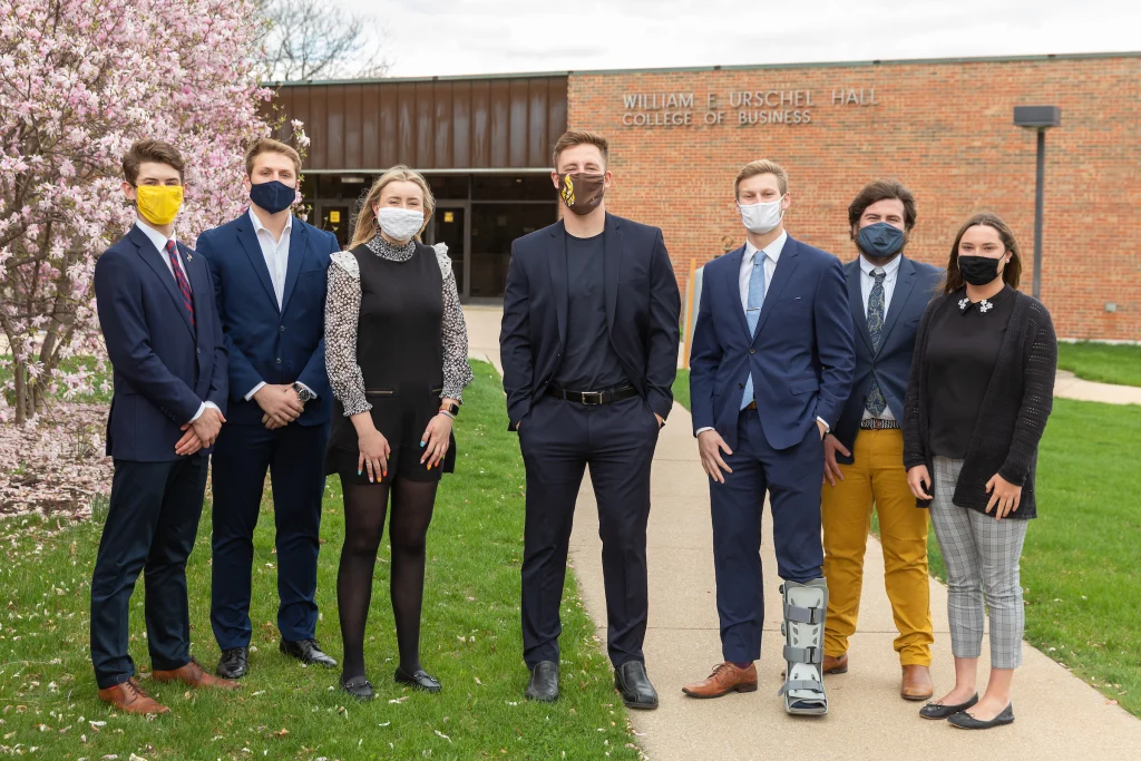Seven people in business attire, wearing masks, stand outside a brick building with spring trees nearby, united in inspiring change within their workplace.