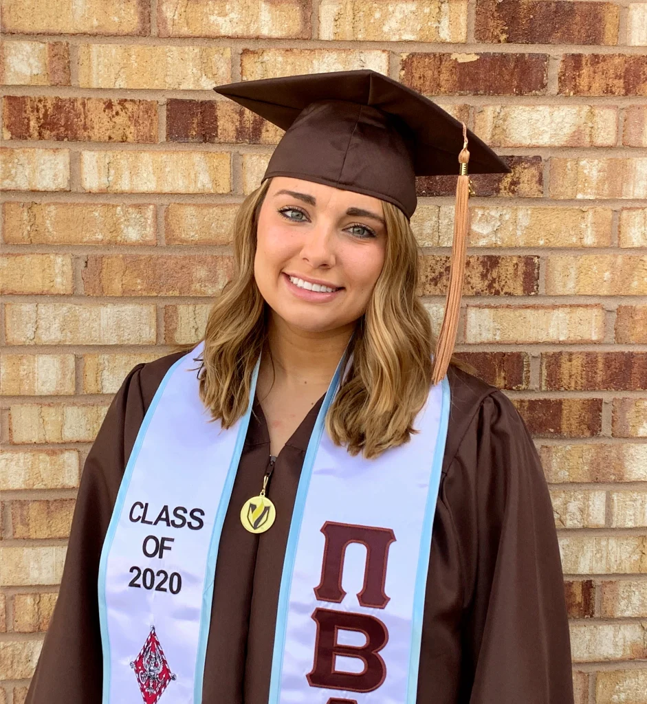 Young woman in a brown graduation cap and gown, smiling in front of a brick wall, Class of 2020 sash visible—proudly forging a new path toward her future.