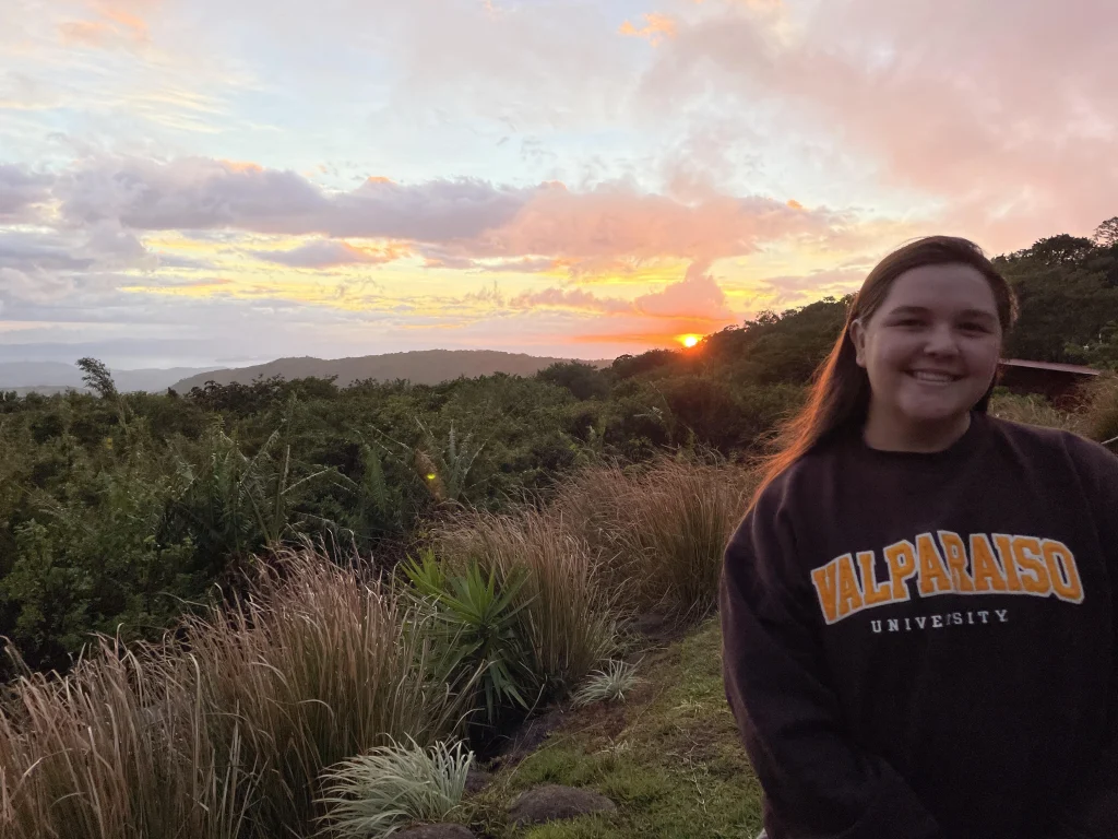 Smiling person in a Valparaiso University sweatshirt stands outdoors at sunset with hills and greenery behind, capturing the spirit of Adventures Abroad and feeling At Home in nature.