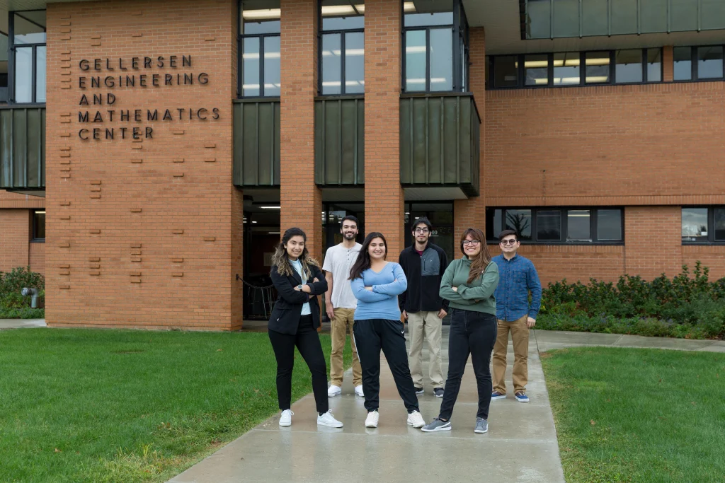 Six people stand smiling outside the Gellerson Engineering and Mathematics Center on a cloudy day, reflecting the spirit of fostering community.