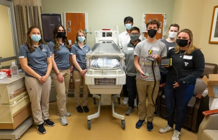 Eight people wearing masks stand around a neonatal incubator in a hospital room, posing for a group photo and showcasing cross-campus teamwork.