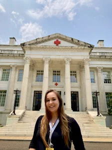 Woman smiling in front of a large white building with columns and a red cross symbol above the entrance, reflecting her commitment to promoting welfare and supporting human welfare initiatives.