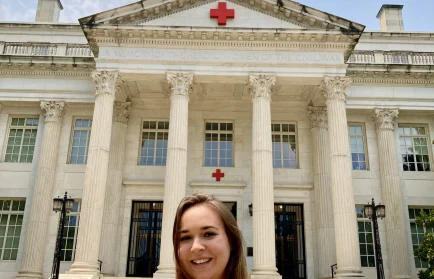 Woman smiling in front of a large white building with columns and a red cross symbol above the entrance, reflecting her commitment to promoting welfare and supporting human welfare initiatives.