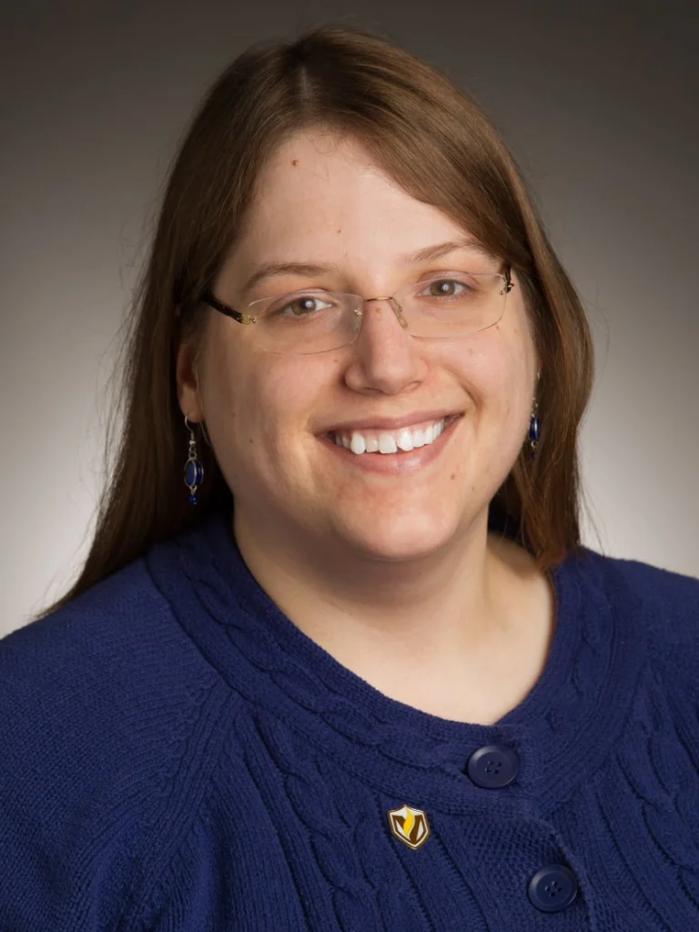 Smiling woman with glasses and long brown hair, wearing a blue sweater and a pin, stands against a neutral background—Executive Director Pudwell of MathPath.