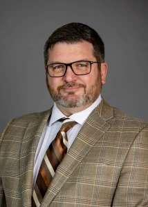 Man with glasses and a beard wearing a plaid suit jacket, white shirt, and striped tie, posing for a portrait as Assistant Vice President at Valparaiso University.