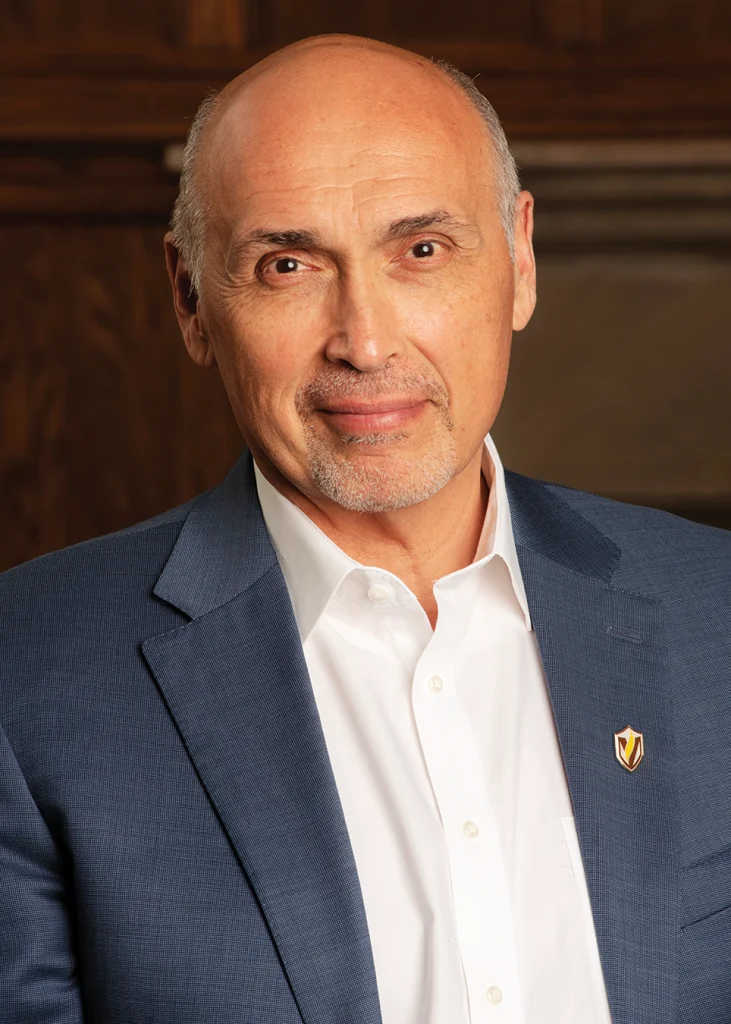 Bald man in a blue suit and white shirt, smiling slightly, with a wooden background—José D. Padilla at Valparaiso University’s President Inauguration.
