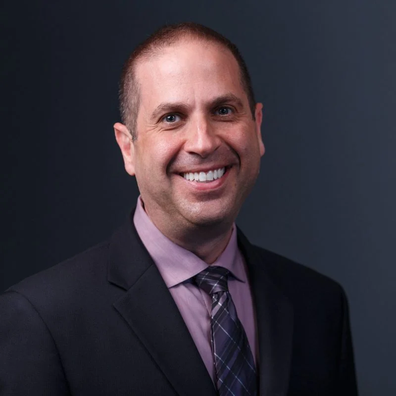 Smiling man in a suit and tie with a dark background, serving as Vice President for Finance and Chief Financial Officer at Valparaiso University.