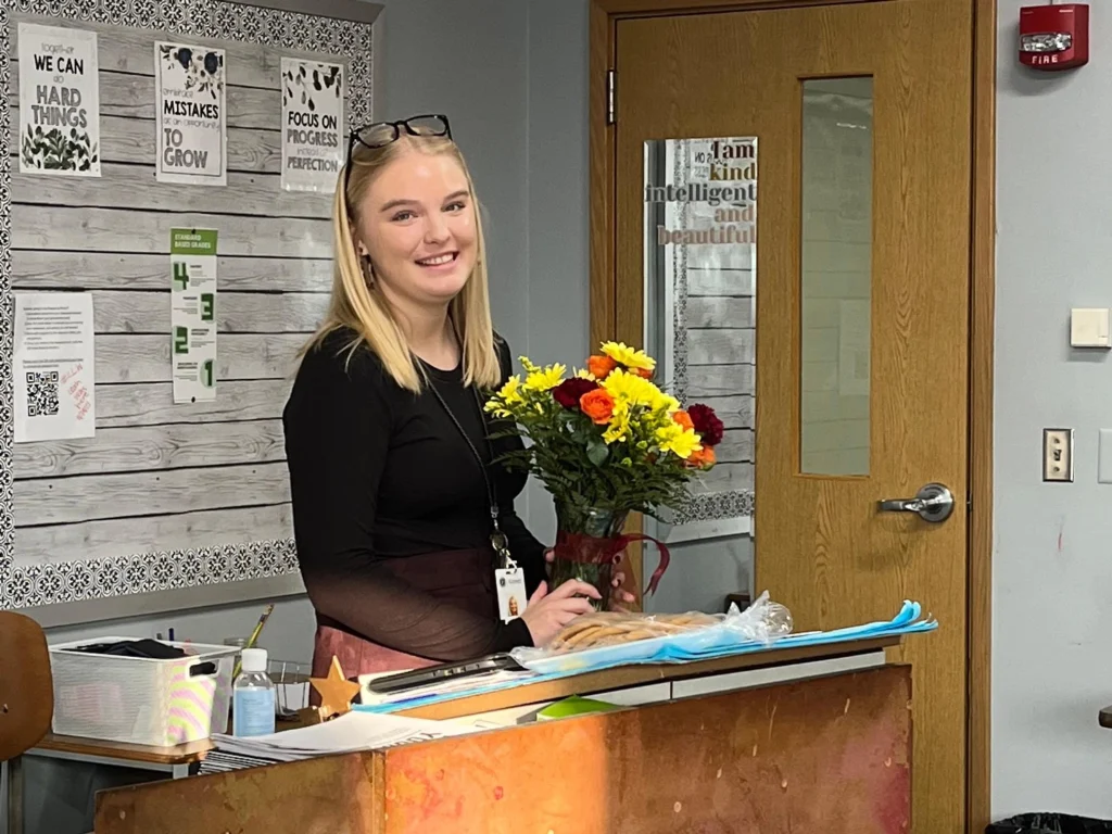 A recognized teacher of the month smiles while holding a bouquet of colorful flowers at her classroom desk.