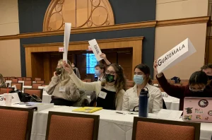 In a Model UN conference room, future leaders raise country name placards, including “Guatemala” and “Israel.”.