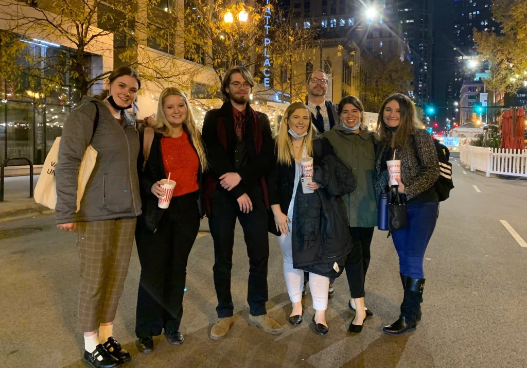 A group of eight future leaders stands smiling on a city street at night, holding drinks and wearing jackets, celebrating new opportunities.