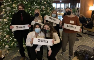 Six people in masks hold "Greece" signs, standing by a decorated Christmas tree in a hotel lobby—future leaders embracing Model UN opportunities.
