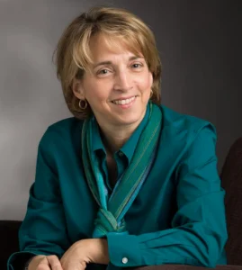 Smiling woman with short hair, wearing a teal shirt and scarf, sits against a gray background—reflecting her dedication to Research and Creative Work at Valparaiso University and her recognition through Awards for Excellence.