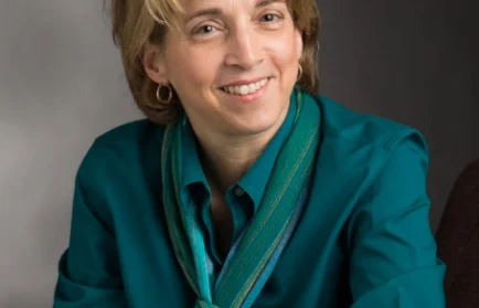 Smiling woman with short hair, wearing a teal shirt and scarf, sits against a gray background—reflecting her dedication to Research and Creative Work at Valparaiso University and her recognition through Awards for Excellence.