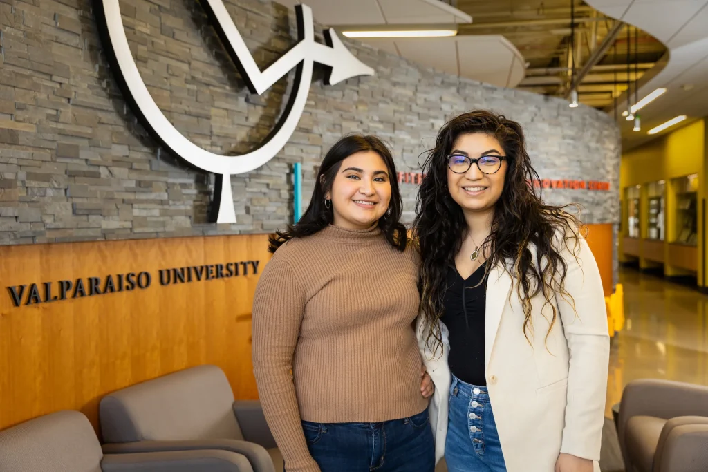 Two women smiling and standing together inside Valparaiso University, reflecting the welcoming spirit of its inclusive community near a large wall logo.