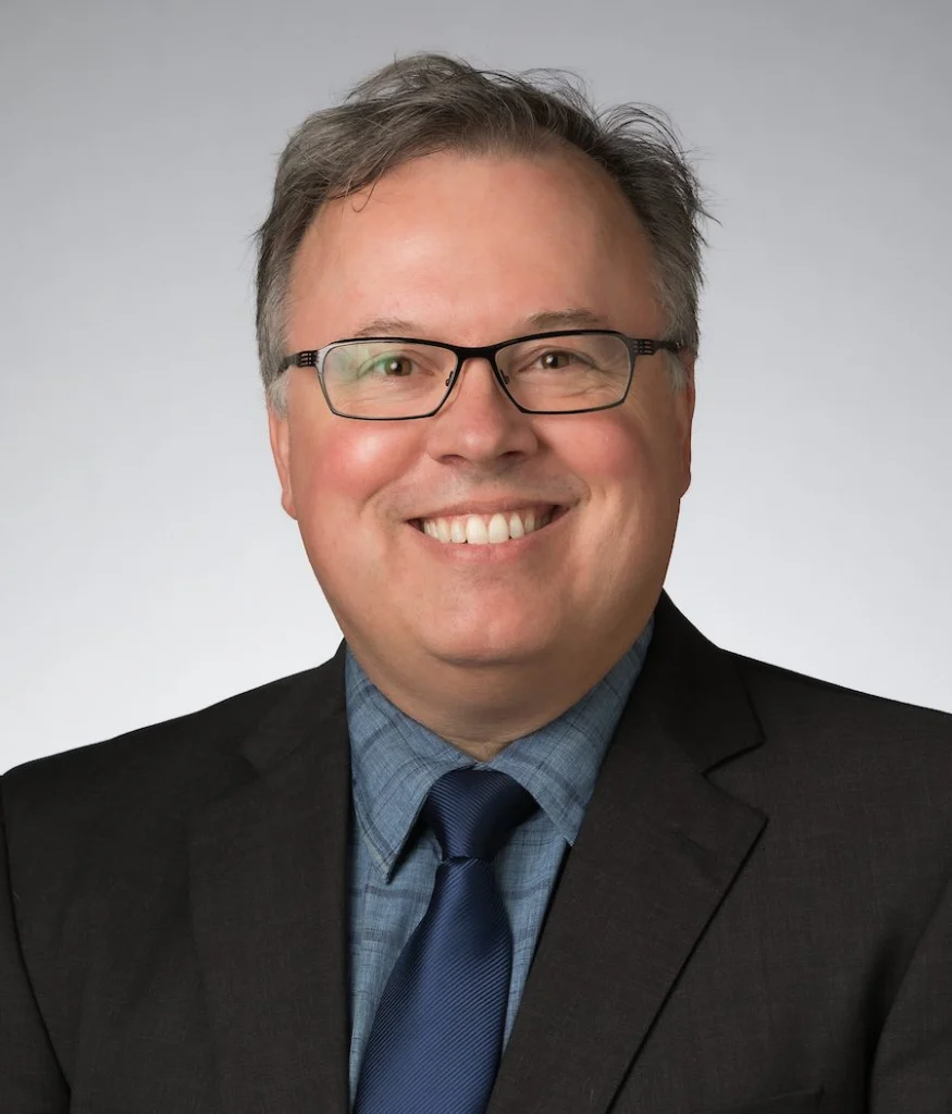 Smiling man in glasses, a dark suit, blue shirt, and tie, stands against a light gray background, representing Valparaiso University student research.