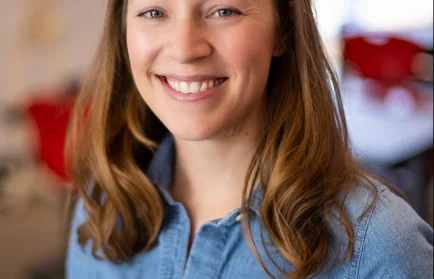 A woman with light brown hair smiles, wearing a blue shirt in a bright, blurred indoor setting—capturing the spirit of Valparaiso University student research and academic enthusiasm.