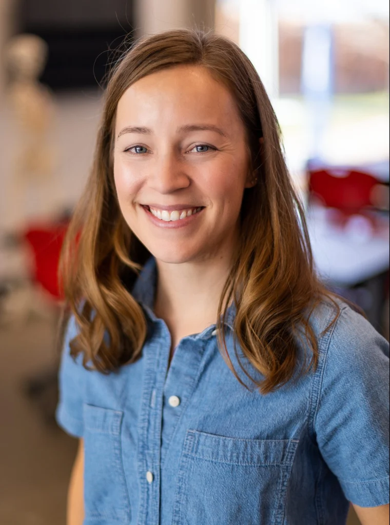 A woman with light brown hair smiles, wearing a blue shirt in a bright, blurred indoor setting—capturing the spirit of Valparaiso University student research and academic enthusiasm.