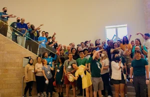 A large, diverse group stands on stairs indoors at Valparaiso University, smiling and pointing at each other in playful poses during a major Mathematics Conference.