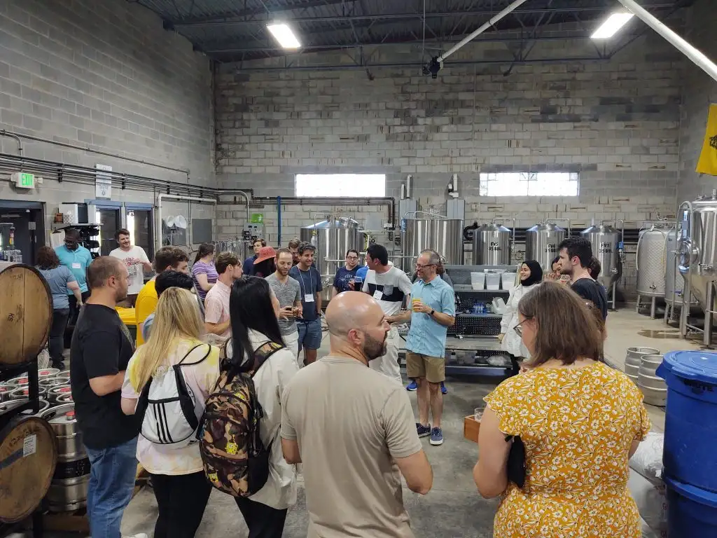 A group of people gather and socialize inside a brewery with metal tanks and barrels in the background, perhaps celebrating after a mathematics conference or an event at Valparaiso University.