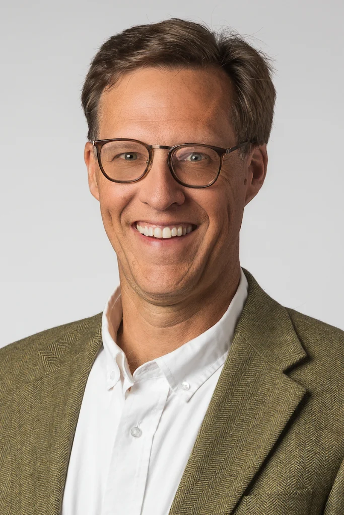 A man wearing glasses, a white shirt, and a green blazer smiles at the camera against a plain background, radiating the confidence that comes from education and a meaningful life.