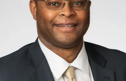 Professor Anton Lewis, in a suit and tie, wearing glasses, smiles at the camera with a plain light background—ready for his next Coffee Break.