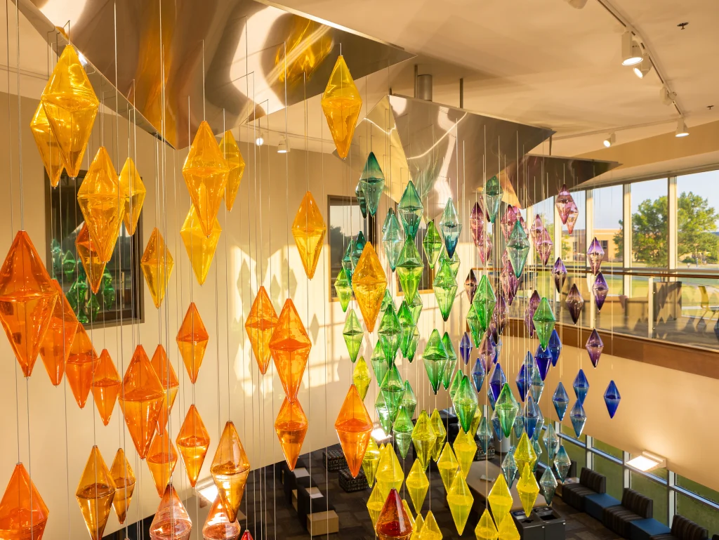 Colorful glass diamonds hang from the ceiling in an art installation at Valparaiso University, crafted using advanced equipment in a sunlit modern building.