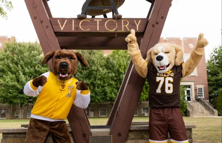 Mascots Beacon and Blaze, dressed in school gear, strike a pose under a large victory bell structure on the college campus.
