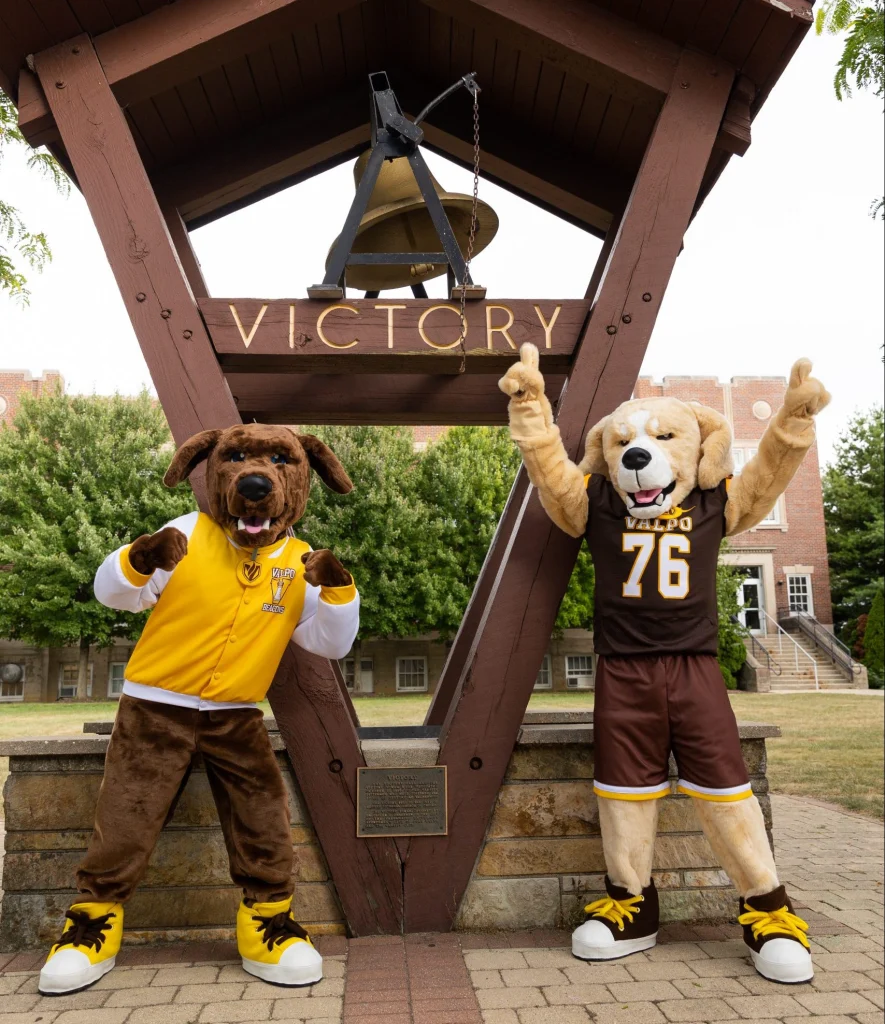 Mascots Beacon and Blaze, dressed in school gear, strike a pose under a large victory bell structure on the college campus.
